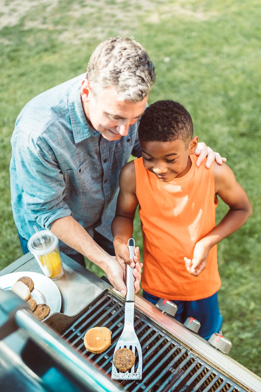 a man and a boy holding a spatula with burger patty on a griller
